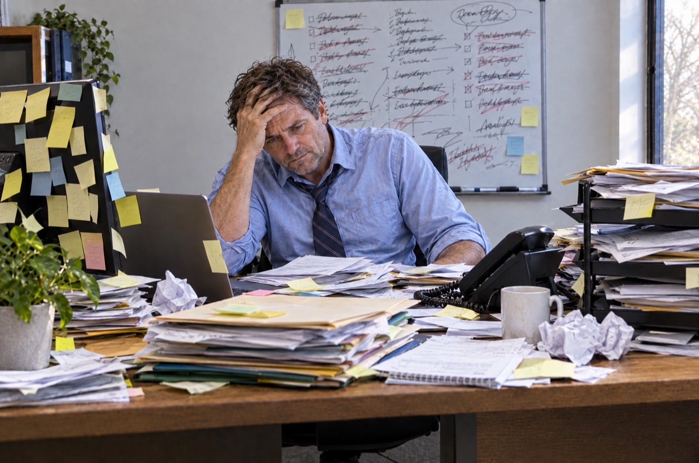 A business owner at his desk buried under stacks of paper, sticky notes and a scribbled-over whiteboard, head in his hand